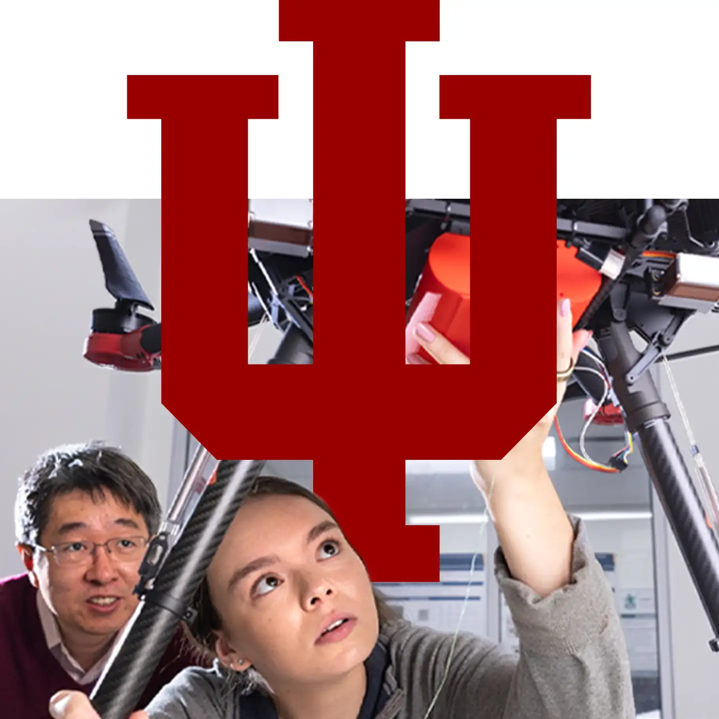 A female student looks up in wonder while working under a large drone, with a male professor watching her intently.