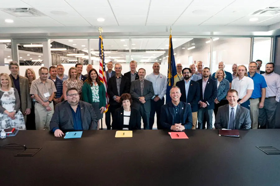 A large group of businesspeople smile in front of American and US flags after signing formal documents laid out on a large table in front of them.